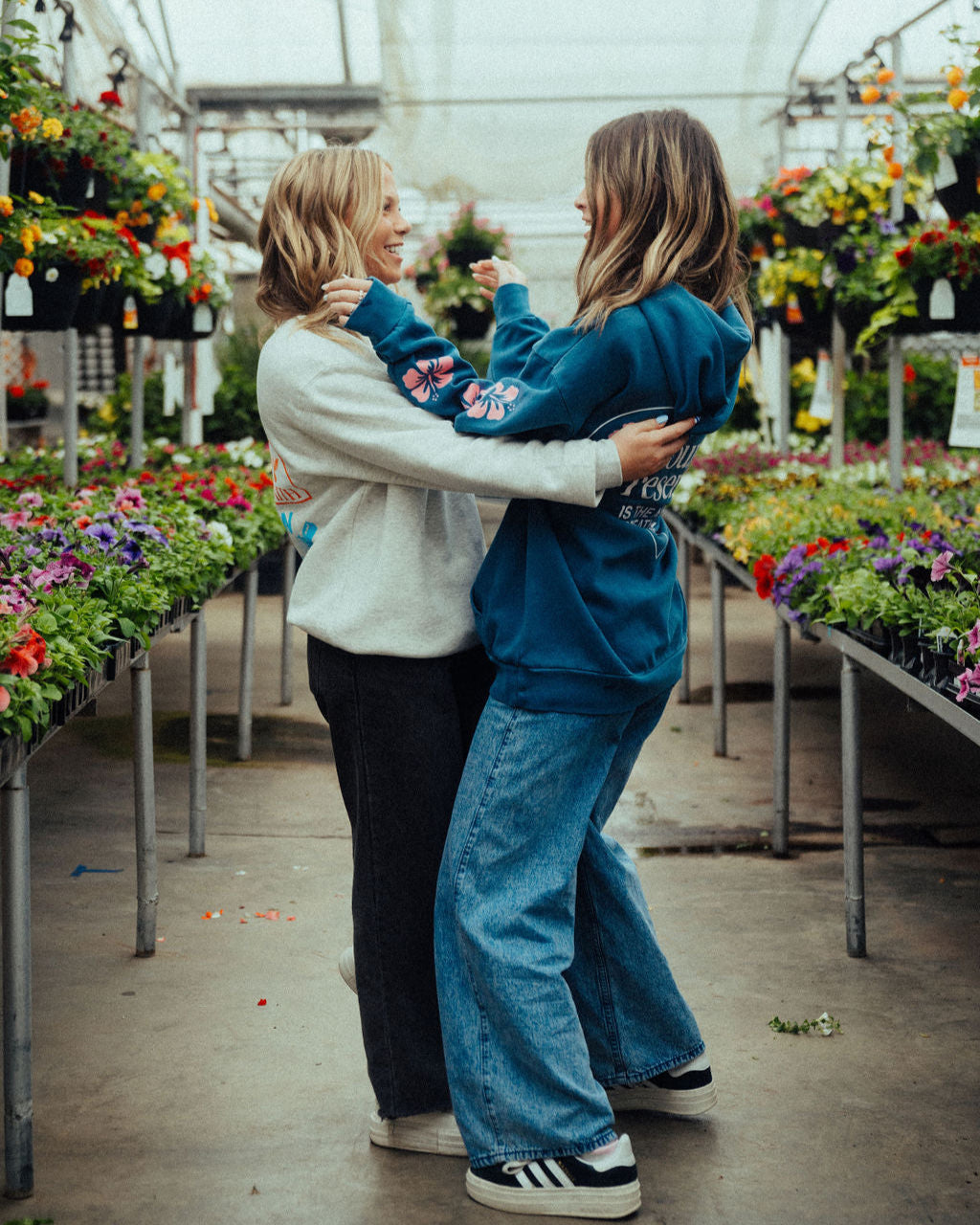 two women hugging in a beautiful greenhouse full of flowers wearing NHIM Apparel christian sweatshirts