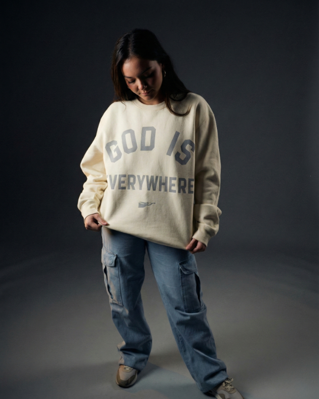 Person wearing a beige sweatshirt with text and blue jeans on a dark background