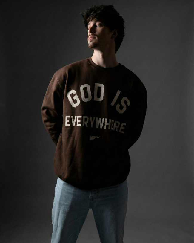 Man wearing a brown sweatshirt with 'GOD IS EVERYWHERE' text on a dark background