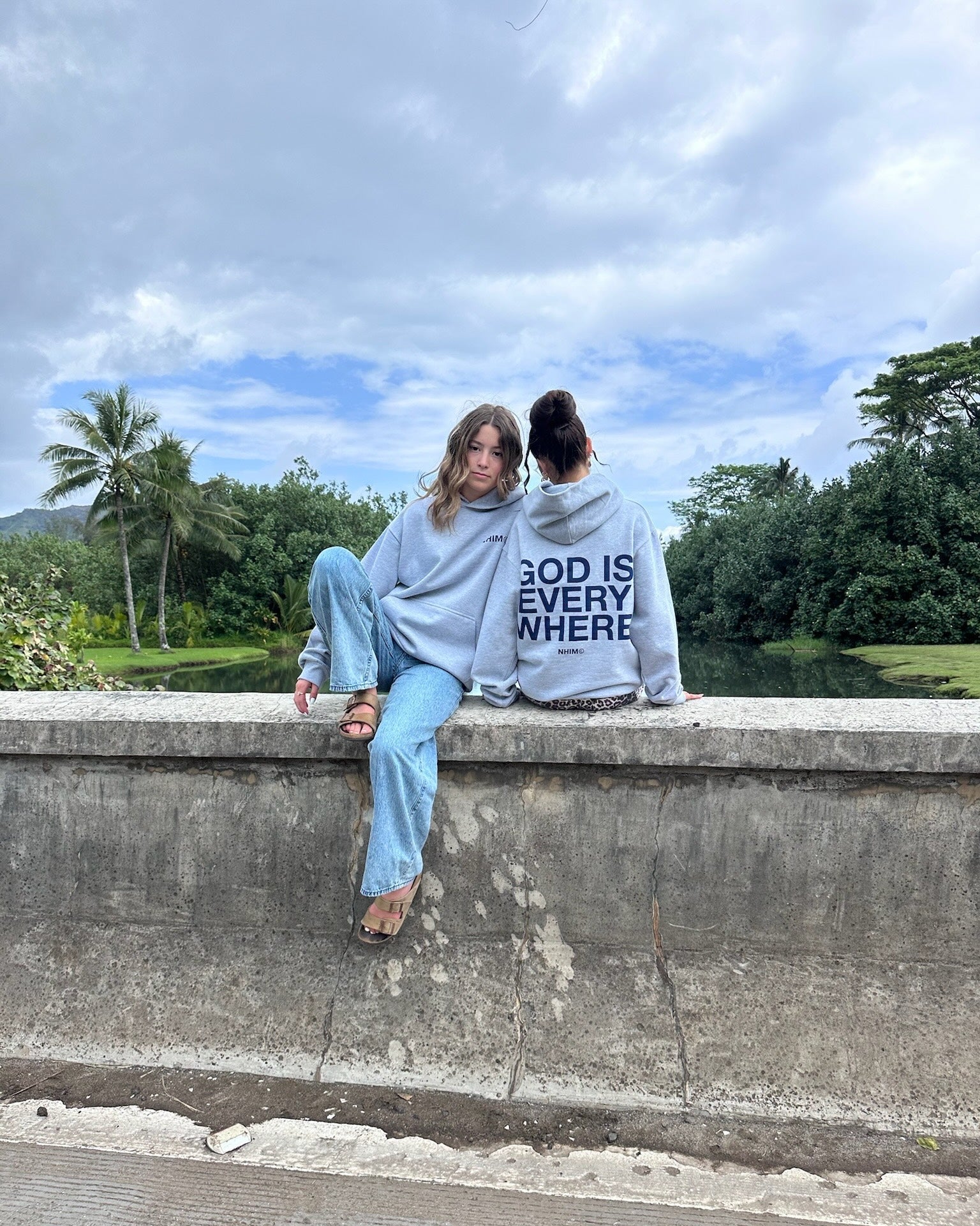two girls sitting on a bridge in kauai wearing God is Everywhere hoodies in grey by NHIM apparel christian clothing brand