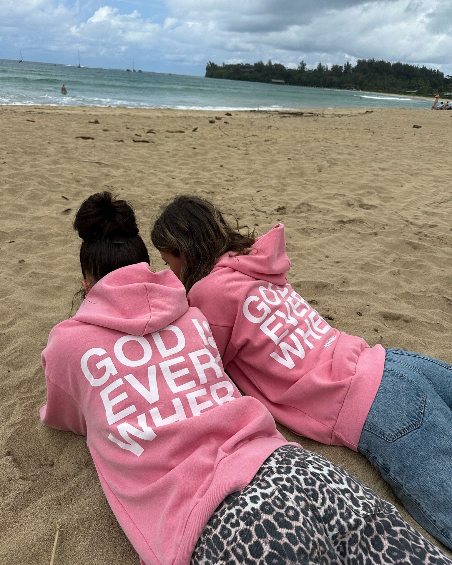 two girls laying on a beach in kauai wearing God is Everywhere hoodies in vintage pink by NHIM apparel christian clothing brand