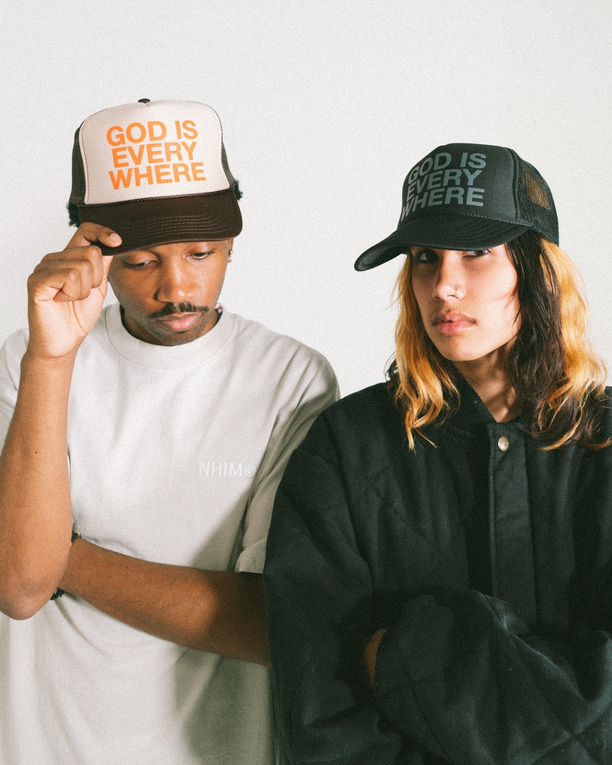GOD IS EVERYWHERE Trucker hat brown and tan with orange writing AND black hat with grey writing. Two models standing next to each other. Christian hat by NHIM APPAREL